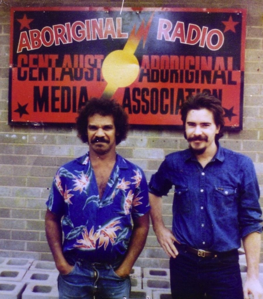 Photograph of two men in blue shirts standing in front of a sign reading Aboriginal Radio..